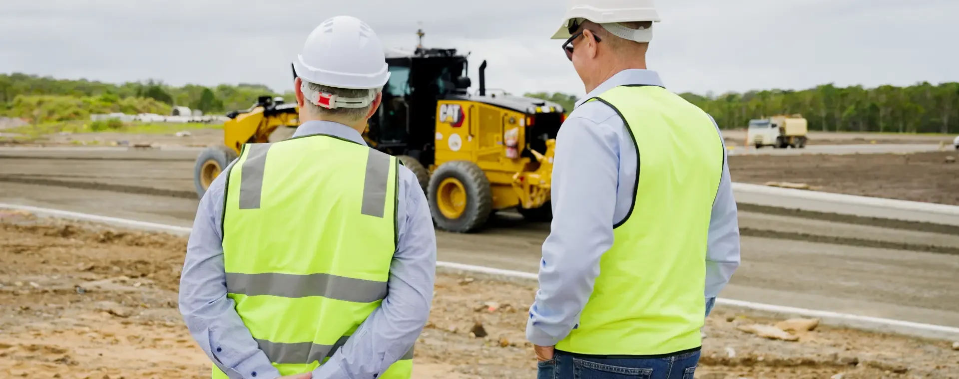 Two people in high-visibility vests and hard hats observe earthmoving works at the Coolum construction site, with a grader in the background.