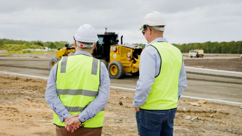 Two people in high-visibility vests and hard hats observe earthmoving works at the Coolum construction site, with a grader in the background.